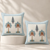 Two decorative square throw pillows with embroidered palm tree motifs and striped blue borders, displayed on a white pedestal against a warm beige background with soft leaf shadows.