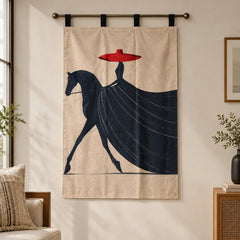 A beige wall hanging or tapestry with a stylized dark navy horse and rider graphic and a bright red hat, suspended from a metal rod in a softly lit room.